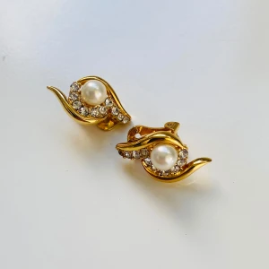 Guldörhängen med pärlor och strass - Säljer ett par eleganta guldörhängen med pärlor och strass. De har en vacker design med en stor pärla i mitten omgiven av gnistrande strass. Örhängena är clip-ons, vilket gör dem perfekta för dig som inte har hål i öronen. De är i nyskick och redo att bäras till fest eller andra speciella tillfällen. Måtten kan ses på bilden med måttbandet.