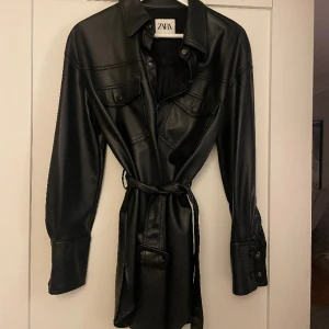 Kavaj i fakeskinn med skärp - Kavaj i fakeskinn material med knytbart bälte i midjan. Helt oanvänd 