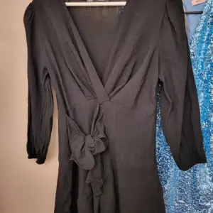 En elegant svart klänning med långärmad design och en knytning i midjan. Klänningen har en v-ringad hals och är tillverkad i ett lätt och bekvämt material. obs vill tilläga att det är en kort klänning 