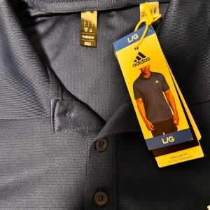 Mörkblå pikétröja från Adidas - En mörkblå pikétröja från Adidas med korta ärmar och tre knappar vid halsen. Tröjan har en broderad Adidas-logga på vänster bröst.