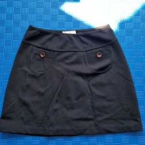 Svart kjol från Kappahl - En svart kjol från Kappahl med två dekorativa knappar framtill. Kjolen är tillverkad av 96% Meryl och 4% Elastan, vilket gör den stretchig när den är våt.