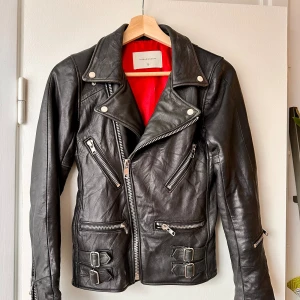 Svart skinnjacka från Deadwood - En svart skinnjacka från Deadwood med röd insida. Jackan har flera dragkedjor och spännen som detaljer. Den har en klassisk bikerstil med en asymmetrisk dragkedja framtill och långa ärmar med dragkedjor vid ärmsluten. Kragen har lite slitage men inget som inte går att fixa med lite läderolja. 