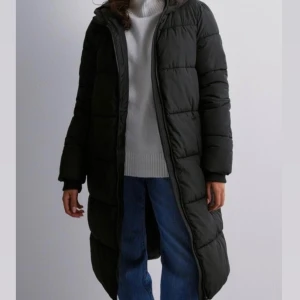 Svart lång dunjacka - En svart lång dunjacka med dragkedja från Nelly.Jackan har långa ärmar och är perfekt för kalla vinterdagar samt för hösten. Använd fåtal gånger och har inga skador. 