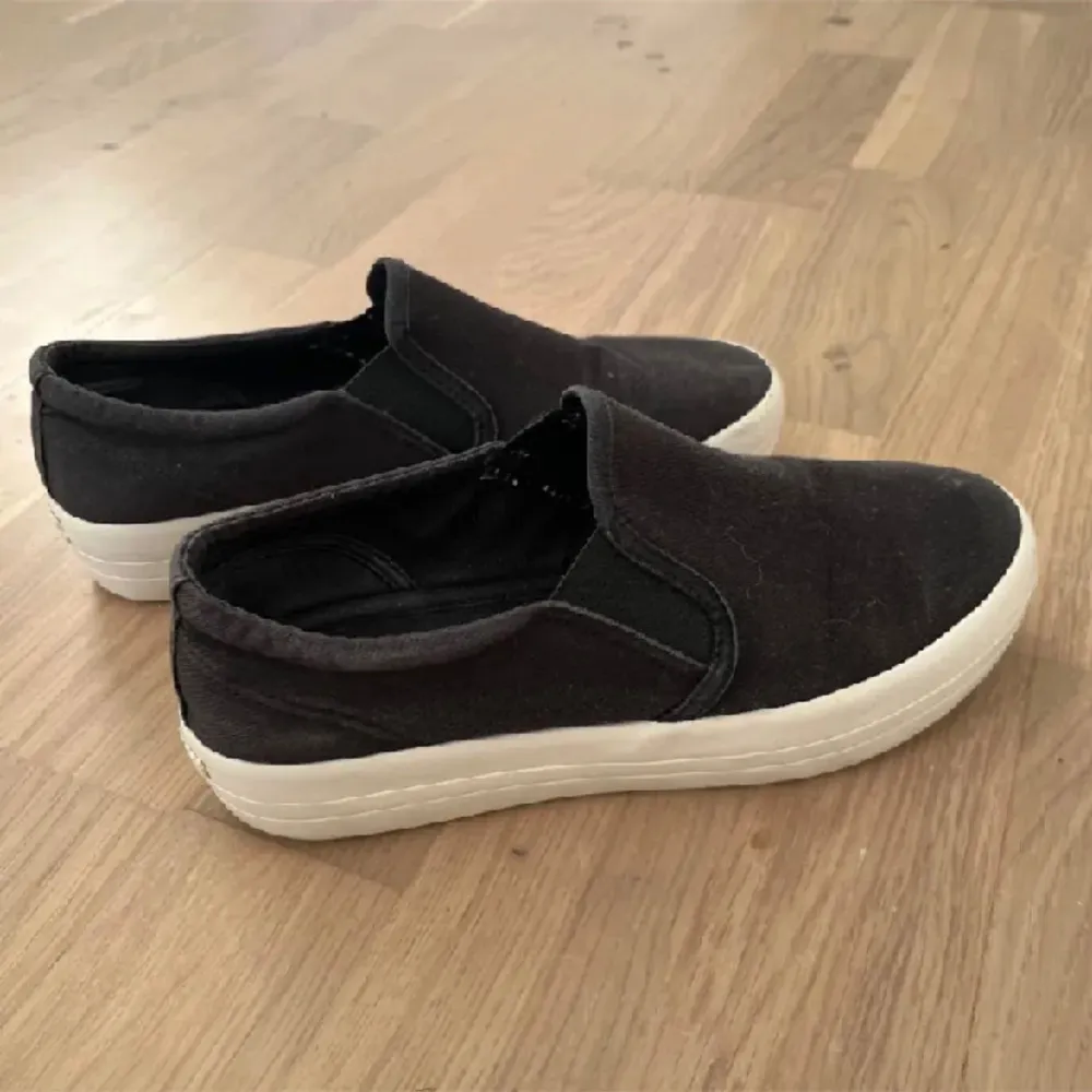 Ett par svarta slip-on skor från Vagabond med vit sula. Skorna har en enkel och stilren design med elastiska paneler på sidorna för enkel på- och avtagning. De är tillverkade i ett syntetiskt material och har en bekväm innersula.. Skor.