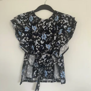 Topp - En fin topp som går att ha på alla årstider förutom vintern. Det finns två band på sidan av tröjan. Jag brukar knyta en fram och en bak til rosetter men där kan du göra som du vill. Jätte fina blommor på tröjan med blåa nyanser. Ärmarna har volanger 