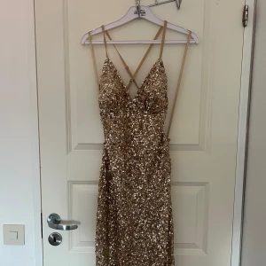 Balklänning från dressroom - Säljer min guldiga balklänning från Dressroom. Klänningen har en helt öppen rygg med justerbara band och är lite längre baktill. Jag är 170 cm och har 5 cm klack på bilden. Den är använd endast en gång och är fortfarande i mycket gott skick, helt ren och fri från fläckar eller smuts. Paljetterna är generellt i mycket bra skick trots att de är känsliga, men några har lossnat längst ner på sidan av klänningen där den släpats mot marken. Nypris: 4 990 kr.  