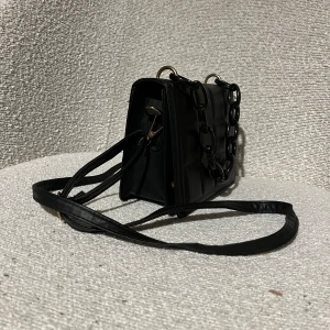 Svart väska - Svart väska i fejkläder med plast handtag som liknar en kedja. Köpt på 2nd hand för 70 kr och jag hittar inget märke. Finns ett svart tunt axelband till 💕 Skriv för fler bilder. Pris kan diskuteras ✉️