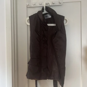 Brun väst från Woman Style - En brun väst från Woman Style med dragkedja och knappar framtill. Västen har en krage och ett bälte i midjan för justerbar passform. Köpt secondhand.