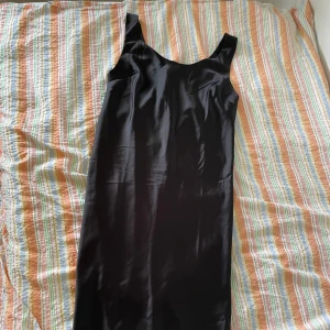 Svart långklänning från H&M - En elegant svart långklänning från ASOS. Klänningen har en enkel och stilren design med breda axelband och är tillverkad i ett glansigt material som ger en lyxig känsla.