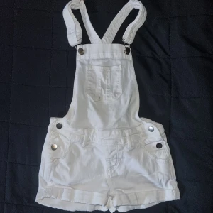 Vita hängselshorts - Ett par vita hängselshorts med justerbara axelband och fickor fram. Shortsen har knappar på sidorna och en ficka på bröstet. Från H&M i storlek 134