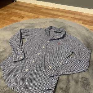 En stilren blå och vit rutig skjorta från Polo Ralph Lauren i storlek S. Skjortan har långa ärmar och knappar framtill. Den är slim fit och har en liten röd broderad logotyp på bröstet.