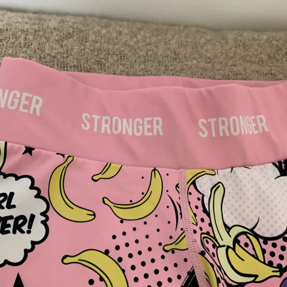 Ett par flerfärgade leggings från Stronger med ett färgglatt serietidningsmönster. Leggingsen har en hög midja och är dekorerade med text och bilder i olika färger som rosa, gul, blå och svart. Nypris 800kr. Jeans & Byxor.