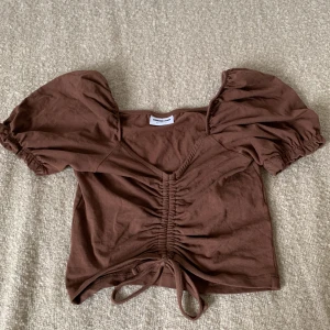 Brun kortärmad topp med rynkdetaljer - En brun kortärmad topp med rynkdetaljer framtill och knytband i nederkant. Toppen har puffärmar med resår vid ärmsluten och en v-ringad halsringning. Toppen är magkort.