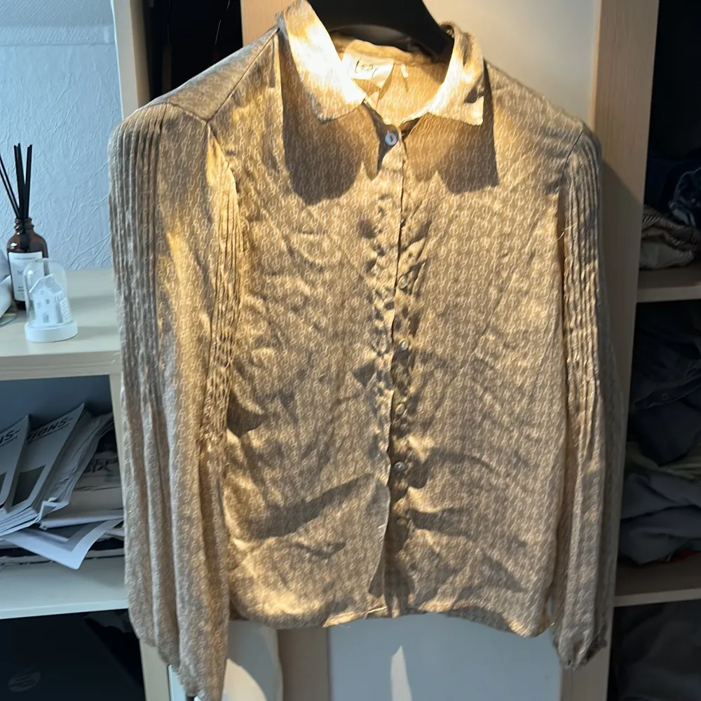 En elegant beige blus från Isay med ett subtilt mönster. Blusen har långa ärmar med veckade detaljer och knappar framtill. Den är tillverkad i ett lätt och bekvämt material, perfekt för både vardag och fest.. Skjortor.