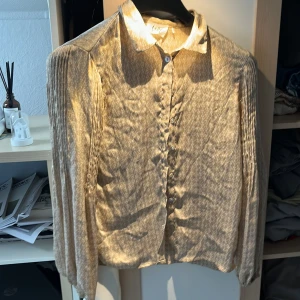 Beige blus från Isay - En elegant beige blus från Isay med ett subtilt mönster. Blusen har långa ärmar med veckade detaljer och knappar framtill. Den är tillverkad i ett lätt och bekvämt material, perfekt för både vardag och fest.