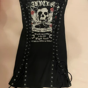 Svart klänning med dödskalletryck och snörning - En svart klänning med ett stort dödskalletryck och text på framsidan. Klänningen har snörning längs sidorna och är ärmlös. Skriv gärna om ni har frågor 💕💕