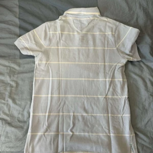 Tommy Hilfiger tröja - Säljer ljusblå Tommy Hilfiger piketröja med vita smala ränder. Perfekt för sommaren och stilren för de som klä sig fint. Storlek S men priset är inte skrivet i sten.