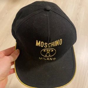 moschino keps - Svartguldig Moschino Milano keps. Bra skick, bara lite dammig! Unisex onesize.  Material: 100% bomull Foder: 100% viskos Tyg: Canvas Skötselråd: Tvättas ej