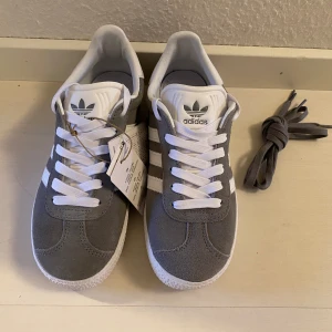 Nya gråa Adidas Gazelle - storlek 36 2/3 (rymliga) - Adidas Originals Gazelle J Färg: Grå (Grey Three-Cloud White-Gold Metallic) Storlek 36 2/3  Nya och oanvända med tags.  Ingår både vita och grå snören.  Populära och slutsålda. 