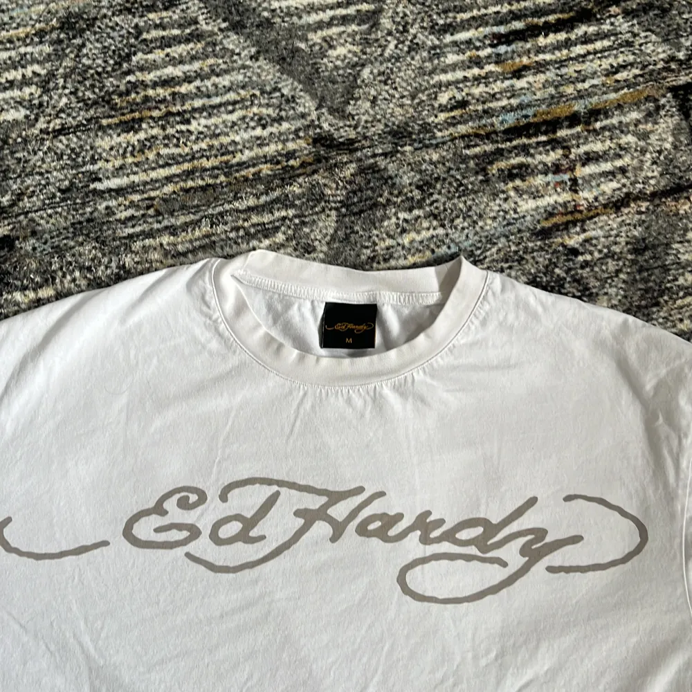 En vit baggy tischa köpt från Ed Hardy, med ett dödskalle motiv på i Ed Hardy stilen i storlek M. Tischan är fläckfri då den endast har använts ett par gånger ca 4 gånger. Den funkar som unisex ifall du gillar den baggy men är avsedd för herr storlek. T-shirts.