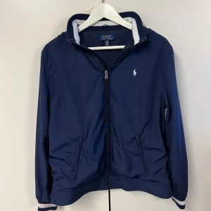 Polo Ralph Lauren Windbreaker - Säljer min riktigt snygga raffe windbreaker! Jackan har Inga slitage och är helt nytvättad.  Jackan är en barnmodell i storlek Xl, vilket motsvarar ungefär vuxenstorleken M. Luvan går att vika in som man ser på sista bilden. Hör av er vid frågor😊🤝 