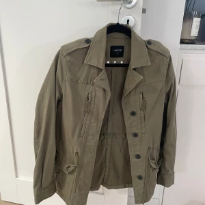 Grön vårjacka  - Militärgrön jacka från Lindex. Väldigt fina detaljer, ex: snörning i midjan och stjärndetaljer. Färgen på jackan är som i de tre första bilder, de två sista är tagna i dåligt ljus(lånade bilder,samma jacka,samma storlek)Pris höjt pga hög efterfrågan!