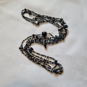 Svart flerlagrat halsband med pärlor - Ett elegant flerlagrat halsband med svarta pärlor i olika former och storlekar. Halsbandet har en mix av små och stora pärlor, inklusive några med mönster. Perfekt för att ge en sofistikerad touch till din outfit.