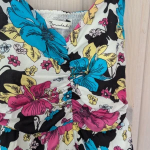 Blommig klänning från Bondelid i 80-tal stil  - En färgglad blommig klänning från Bondelid med breda axelband och en åtsittande överdel. Klänningen har ett mönster med stora blommor i blått, rosa, gult och svart på en vit bakgrund. Tillverkad av 100% bomull.strl S I i mycket fint skick 