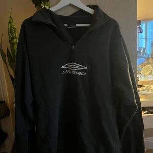 Svart tröja från Umbro - En svart tröja från Umbro med en dragkedja vid halsen och Umbro-logotypen tryckt på bröstet. Tröjan har långa ärmar och en hög krage.