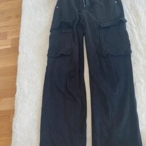 Svarta cargobyxor från Bershka - Svarta cargobyxor från Bershka i storlek EUR 36. Byxorna har flera fickor med knappar och en dragkedja framtill. De är av modellen 'Medium Rise Cargo Straight'.