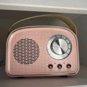 Rosa retroinspirerad högtalare - En oanvänt rosa retroinspirerad högtalare med en rund volymknapp och tre knappar för kontroll av uppspelning. Högtalaren har en beige bärrem. Den har flera anslutningsmöjligheter inklusive Type-C, TF-kort, USB och AUX.