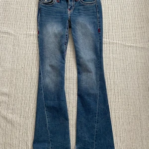 True religion jeans - Blå jeansbyxor från True Religion med låg midja och utsvängda ben. Byxorna har dekorativa sömmar i rött och blått samt fickor med knappar på baksidan. De är uppsydda 