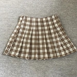 Brun rutig kjol - En veckad kort kjol som passar perfekt till hösten. Kjolen är endast använd ett fåtal gånger och har inga defekter. Den är vit och brun rutig. 