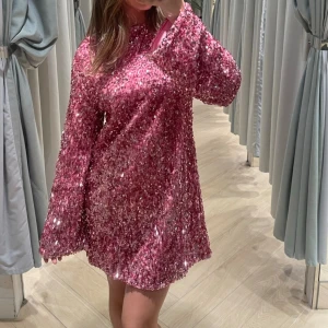 Rosa paljettklänning med rosett - En glittrig rosa klänning täckt med paljetter. Klänningen har långa ärmar och en stor rosett på baksidan.