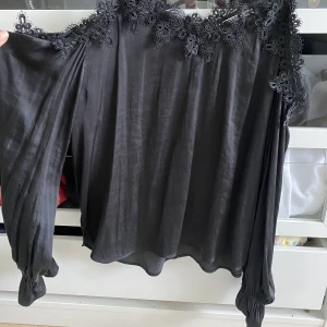 Svart blus med spetsdetaljer - Fint skick, men använd. En svart topp med satin-känsla. Offshoulder och med spetsdetaljer.