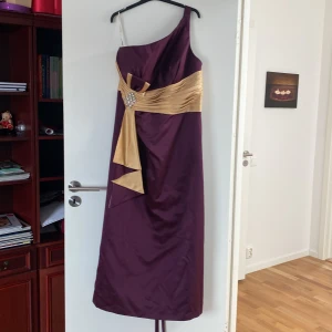 Fantastiskt festklänning  - Lång elegant festklänning. Snygg klänning i mörk lila färg med fin detalj i guld och strass. Snörning bakom i ryggen gör det att justera storleken på överdelen. Använd endast en gång på ett bröllop. Storlek L/XL. Finns även en exakt likadan i barnstorlek 