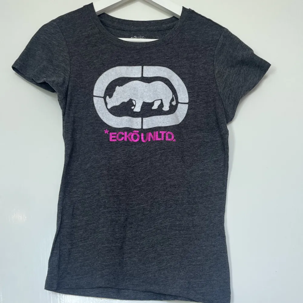 Grå T-shirt från Ecko med motiv av en vit noshörning och rosa/lila text som läser *ECKÖ UNLTD. Fint skick. Jag är S och 175cm lång.. T-shirts.