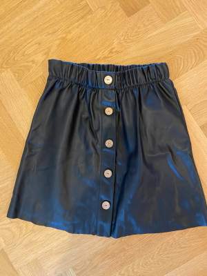 Kort kjol i skinnimitation med knappar, slutar ovanför knät 