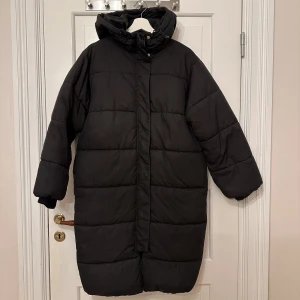 Lång pufferjacka  - Svart lång pufferjacka från H&M i storlek XS. Oversized, rymlig på mig som vanligtvis har M men inte för stor. Fickor på sidan. Luva och tvåvägsdragkedja. Resår nedtill så den är puffig” och inte rak. Går nedanför knäna på mig som är 160. Använd fåtal ggr