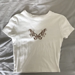 Vit t-shirt med brunt tryck - En jättefin vit croppad t-shirt med ett brunt fjärils tryck på. Säljer denna då den inte är min stil längre. Jättefint skick och har inte används många gånger.