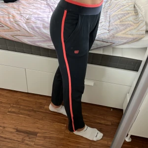 Wilson överdragsbyxor träning - Mörkgrå överdragsbyxor till träning från Wilson. Med cool orange kontrastfärg på sidorna och i midjan och har fickor. Har även blixtlås nedtill byxbenen som kan dras upp för att ta av byxorna över skorna. Storlek XS.