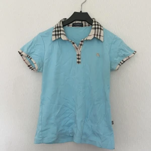 Vintage burberry piké/polotröja - En ljusblå burberry piké/polotröja med en knapp samt burberry mönster på kragen och i ärmarna. Gör fin färg och gullig topp men får aldrig användning av den.