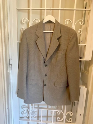 Ljusgrå retrokavaj 90-tal – Joyboy - Retrokavaj från Joyboy. Smårutigt textilmönster, ljusgrått med mörkare inslag. Fint skick.  Storlek: 150 63 % ull. 23 % polyester. 8 % polyamid. 6 % viskos. Foder: 100 % acetat.