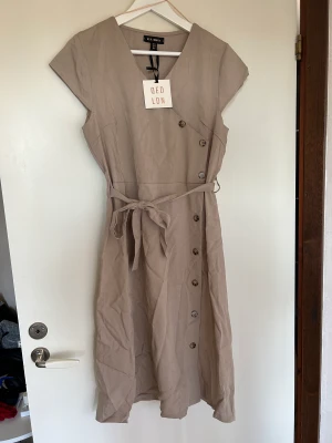 Oanvänd klänning - En brun-beige klänning från BUBBLEROOM. Knappar som går att öppna och ett band i midjan. Prislappen sitter kvar då klänningen är oanvänd. Längden är precis under knäna för mig som är 175 cm lång. 