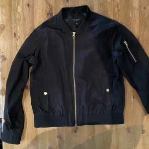 Bomberjacka - Jacka med ficka på vänster arm och två vanliga fickor som stängs med en knapp. Dragkedjorna och knapparna är mässingfärgade.