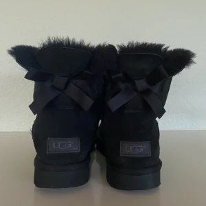 Svarta Uggs (mini bailey bow) i storlek 39 - Ett par svarta ”mini bailey bow” från märket UGG i storlek 39. Köpta i en UGG-butik i USA för några år sedan. Skorna har vissa tecken på användning men i övrigt i bra skick! Skriv gärna för fler bilder, så kan jag visa ”defekterna” <3