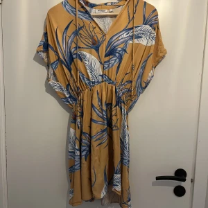 Gulblå sommarklänning (Gail dress från Weekend), storlek XS - Kort sommarklänning med blått mönster, i tunn viskos. V-ringning och snöre som går att knyta i halsen. Resår i midjan. Jag har sytt ett par cm framtill där sömmen gått upp, se bild 4.  Hund finns i hemmet, men jag tvättar klänningen innan den skickas.