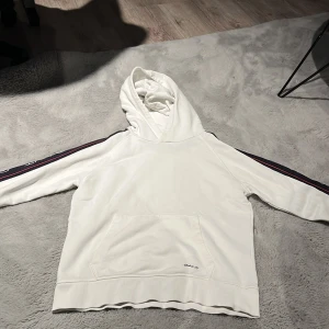 Vit hoodie från GANT med ränder - Säljer en vit hoodie från GANT med huva och känguruficka. Ärmarna har snygga marinblå och röda ränder längs sidorna och GANT-loggan är broderad framtill. Perfekt för en chill och sportig look.Den var några fläckar men de går att ta bort. 