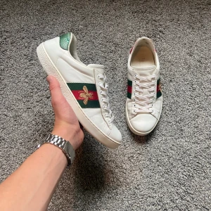 Gucci Ace vita sneakers med bi - Gucci Ace sneakers i vitt skinn med klassiska gröna och röda ränder på sidorna och broderad guldfärgad bi. Grön och röd detalj i metallic på hälen. Platt sula och snörning framtill. Snygg och ikonisk modell för dig som gillar lyxiga detaljer.