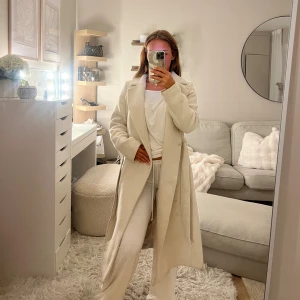 Beige kappa med bälte från H&M - Snygg beige kappa från H&M med klassisk krage och bälte i midjan. Använd ett antal gånger och har därav någon fläck🌸